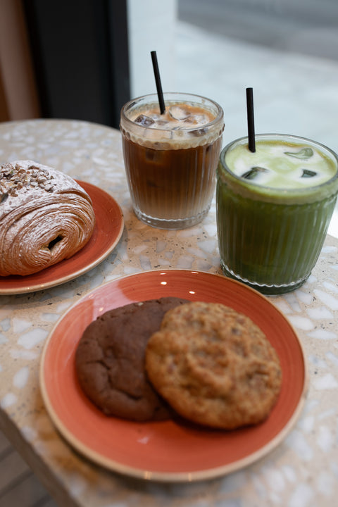 Two cookies on a plate, croissant, one cup of iced matcha, and iced coffee