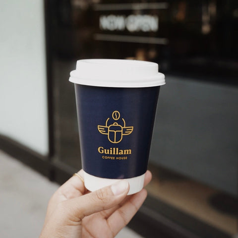 Hand holding a blue coffee cup with 'Guillam Coffee House' logo in a blurred cafe setting
